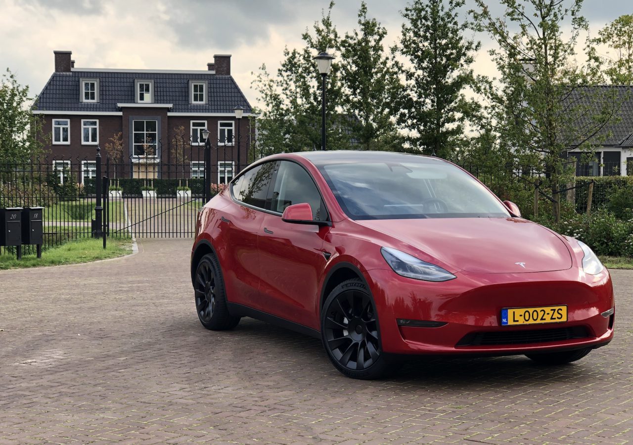 Een rode Tesla Model Y staat geparkeerd op een geplaveide oprit voor een afgesloten woonwijk met bakstenen huizen en bomen, wat de elegantie van het Tesla Model laat zien.