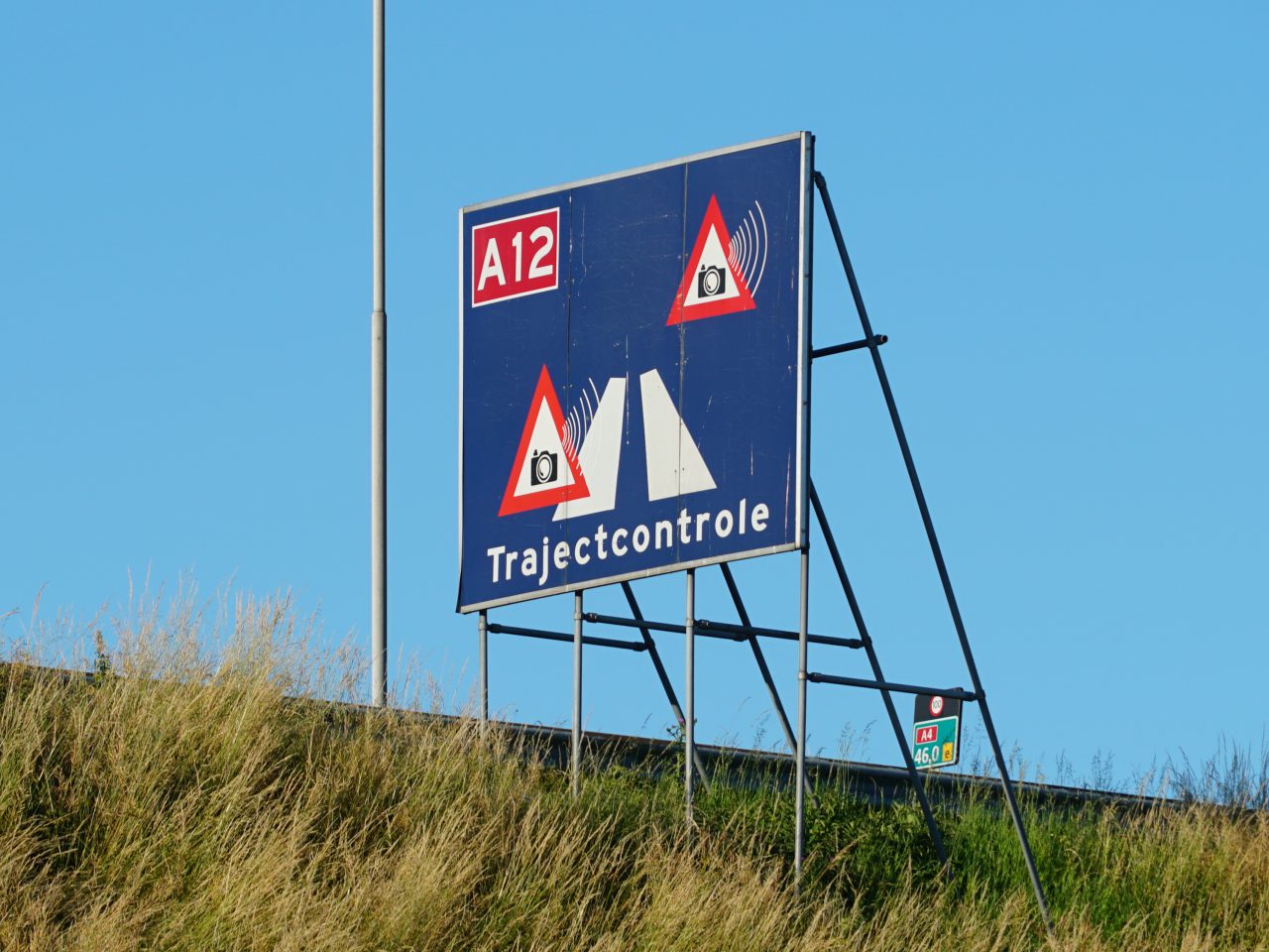 Een blauw bord met daarop 'A12 Trajectcontrole' dat snelheidshandhaving op de A12 aangeeft, voorzien van flitspaalpictogrammen en een rood driehoekswaarschuwingssymbool, herinnert automobilisten aan mogelijke verkeersboetes 2022 voor snelheidsovertredingen.