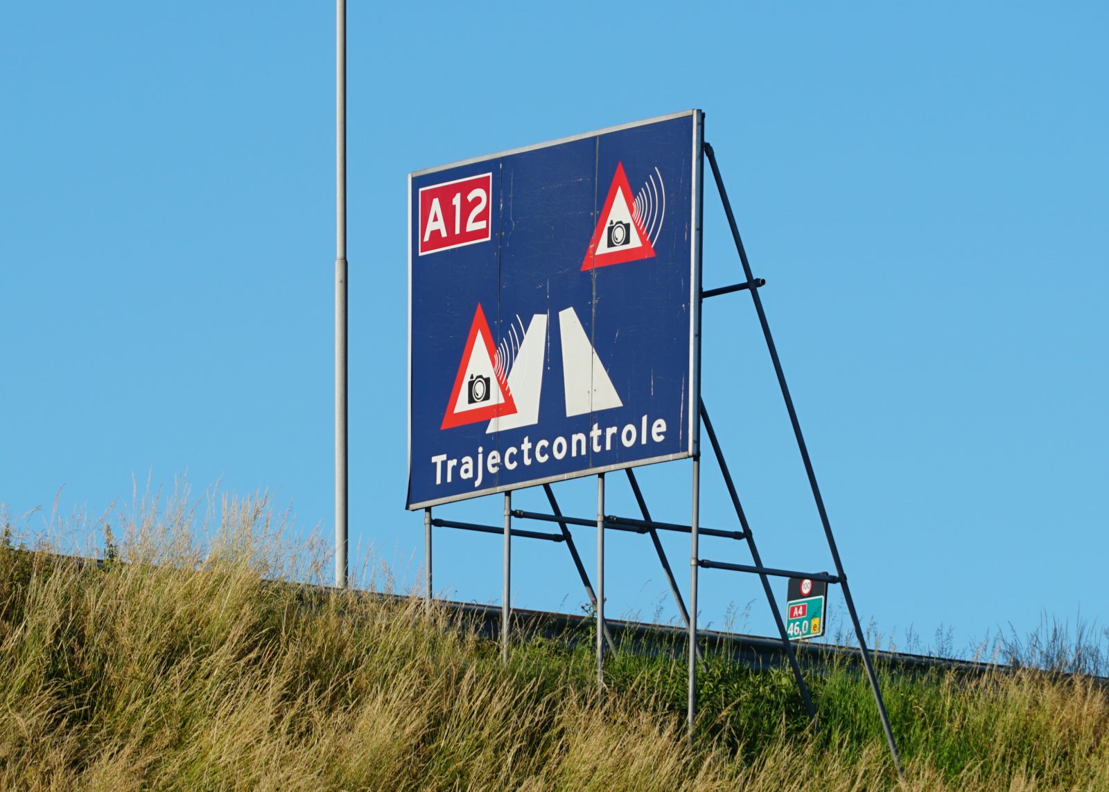 Een blauw bord met daarop 'A12 Trajectcontrole' dat snelheidshandhaving op de A12 aangeeft, voorzien van flitspaalpictogrammen en een rood driehoekswaarschuwingssymbool, herinnert automobilisten aan mogelijke verkeersboetes 2022 voor snelheidsovertredingen.