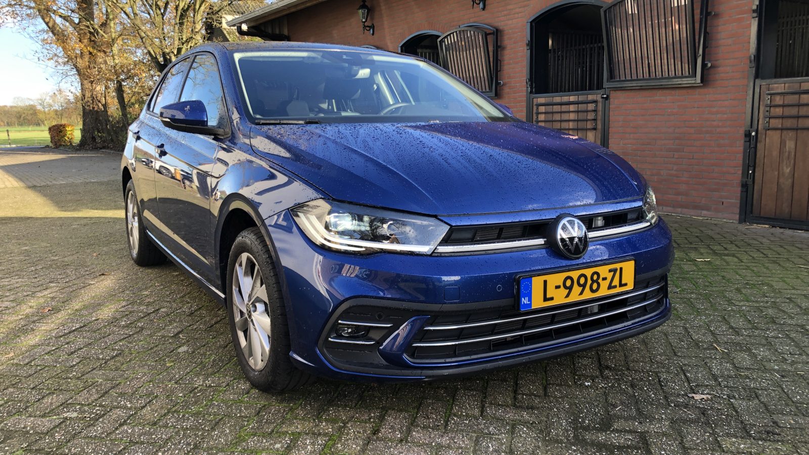 Een blauwe Volkswagen Polo met kenteken L-998-ZL staat geparkeerd op een geplaveide oprit voor een bakstenen gebouw met houten deuren.