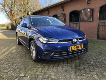 Een blauwe Volkswagen Polo met kenteken L-998-ZL staat geparkeerd op een geplaveide oprit voor een bakstenen gebouw met houten deuren.