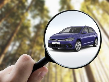 Een hand houdt een vergrootglas vast en focust op een blauwe Volkswagen Polo, bekend om zijn betrouwbaarheid onder betrouwbare auto's, met een onscherpe achtergrond van hoge bomen.