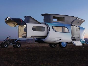 Een futuristische, zilverblauwe Whale Trailer Cabin met verlengde zijpanelen en bovendelen, geparkeerd op grasachtig terrein in de schemering, met een terreinwagen ernaast.