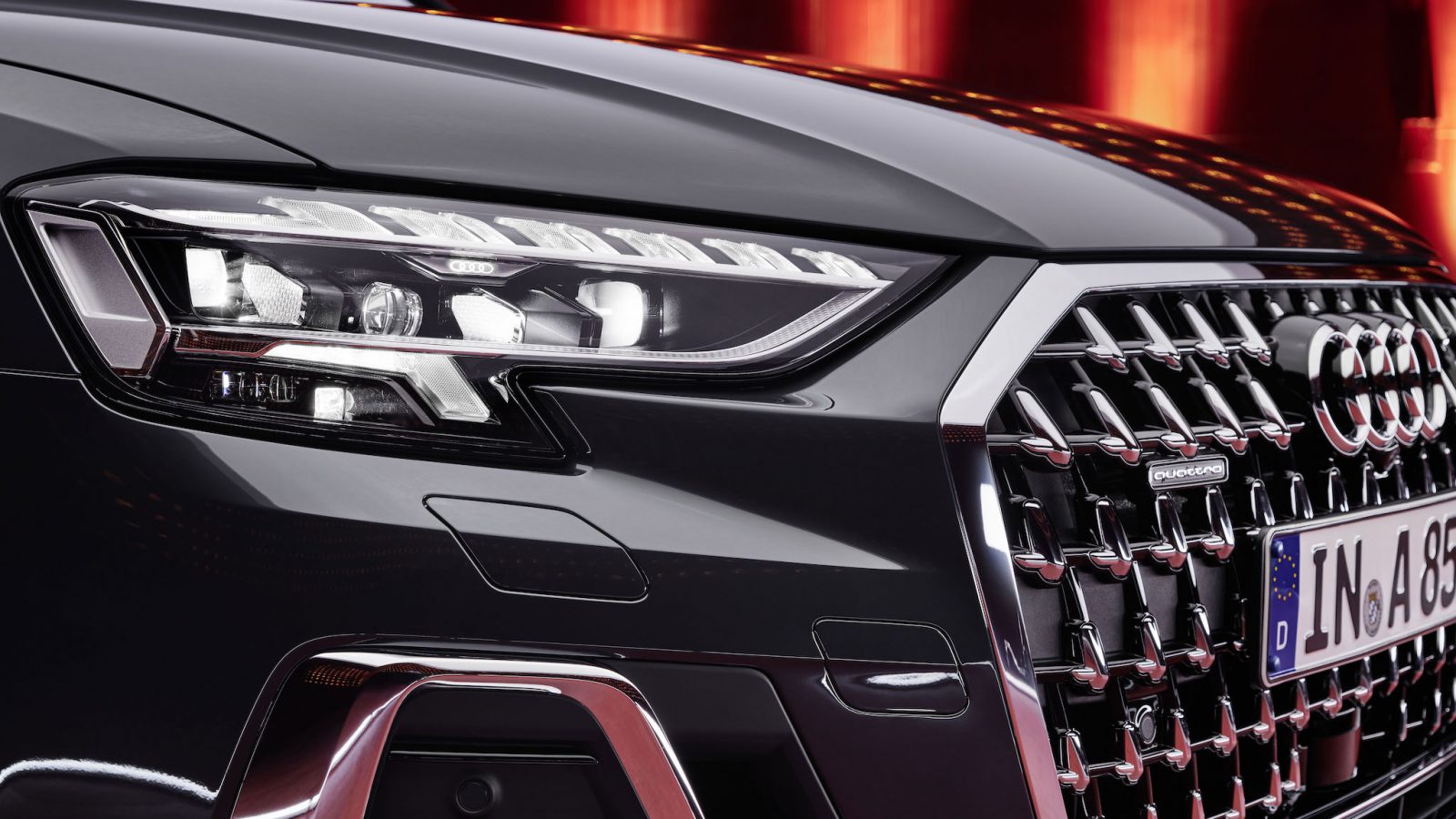 Close-up van de voorkant van een zwarte Audi A8 luxe sedan met een opvallende grille en strakke LED-koplampen, tegen een achtergrond van levendige rode lichten.