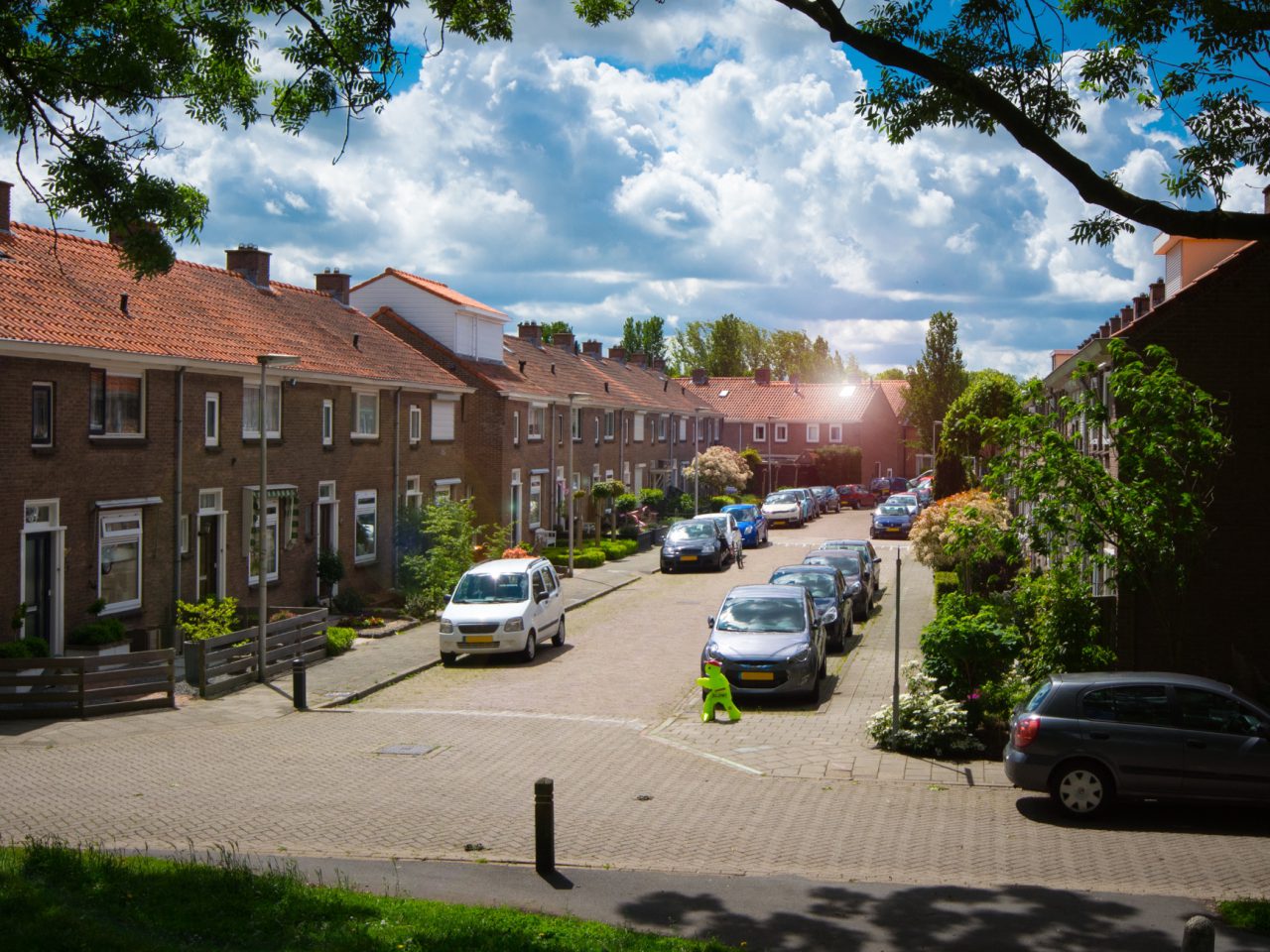 Een zonnige woonstraat met geparkeerde auto's, bakstenen rijtjeshuizen en een klein centraal grasveld met een plastic stoel. Bomen en groen omringen het gebied onder een gedeeltelijk bewolkte hemel, waar af en toe de CO2-uitstoot van auto's de vredige sfeer nauwelijks verstoort.