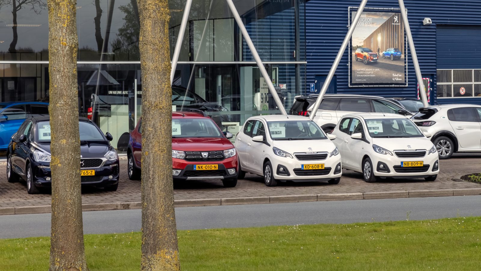 Een rij geparkeerde auto's met te koop-borden voor een dealergebouw met glazen wanden op een gedeeltelijk bewolkte dag, gezien van achter twee bomen, weerspiegelt de optimistische verkoopprognose auto 2022.