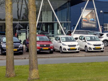Een rij geparkeerde auto's met te koop-borden voor een dealergebouw met glazen wanden op een gedeeltelijk bewolkte dag, gezien van achter twee bomen, weerspiegelt de optimistische verkoopprognose auto 2022.