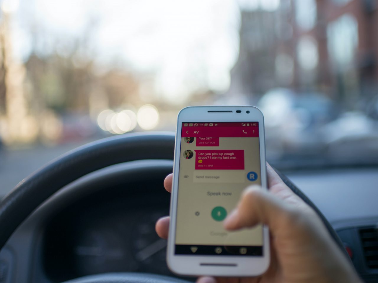 Een hand houdt een smartphone vast met een berichten-app open terwijl hij zich boven het stuur van een auto bevindt, wat aangeeft dat de gebruiker aan het sms'en is tijdens het rijden. Overweeg om een van de 4 apps tegen afleiding in het verkeer te gebruiken om gefocust en veilig onderweg te blijven.