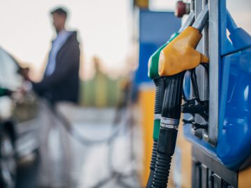 Close-up van het mondstuk van een benzinebrandstofpomp bij een benzinestation met op de achtergrond een persoon die zijn auto tankt.