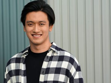 Een man met kort, donker haar, gekleed in een zwart-wit geruit overhemd over een zwart T-shirt, glimlacht naar de camera voor een wand van golfplaten, wat doet denken aan de zelfverzekerde glimlach die Guanyu Zhou zou kunnen hebben als hij in 2022 voor Alfa Romeo rijdt. de F1.
