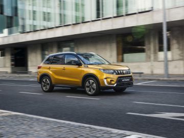 Een gele Suzuki Vitara rijdt door een lege stadsstraat met moderne glazen gebouwen op de achtergrond.