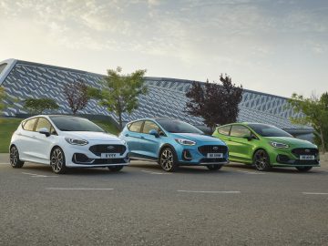 Drie Ford-auto's, waaronder een strakke Ford Fiesta, wit, blauw en groen gekleurd, staan op een heldere dag op een rij geparkeerd voor een modern gebouw.