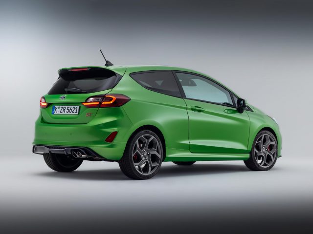 Een groene, compacte tweedeurs Ford Fiesta wordt rechtsachter getoond, met glanzende lichtmetalen velgen, een achterspoiler en dubbele uitlaatpijpen. De achtergrond is een effen verloop. De auto heeft een Europees kenteken.