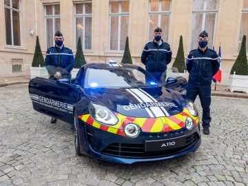 Drie agenten in uniform van de Franse politie staan naast een donkerblauwe auto van de gendarmerie met rode en gele punthaken, geparkeerd op een geplaveide ondergrond voor een gebouw met meerdere grote ramen.