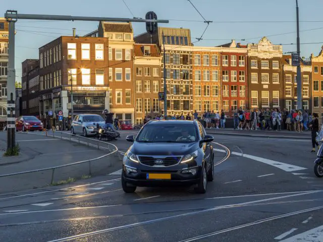 Een drukke stadsstraat met verschillende auto's en motorfietsen die over de bochtige weg navigeren, waar gebouwen op de achtergrond staan. Een menigte mensen verzamelt zich op het trottoir en borden geven de maximale snelheid weer om een vlotte verkeersstroom in deze bruisende steden te garanderen.