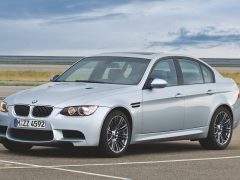 Een zilveren BMW M3 sedan, één van de Top 5 auto's voor auto-investeringen, staat geparkeerd op een leeg verhard terrein met een bewolkte lucht op de achtergrond.