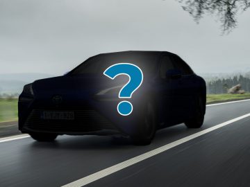 Afbeelding van een auto die over een lege weg rijdt, met een groot vraagteken op de voorkant. De achtergrond is voorzien van een bewolkte lucht en bomen, perfect voor een automatische beoordeling waarbij je je afvraagt of dit voertuig de juiste keuze is voor je volgende avontuur.