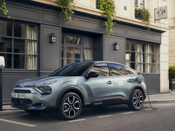 Citroën ë-C4 geparkeerd in een stadsstraat, aangesloten op een laadstation. Het gebouw op de achtergrond heeft grote ramen met gordijnen en overhangende groene planten. Mis dit niet: reserveer uw model vandaag nog om te profiteren van de beschikbare subsidies.