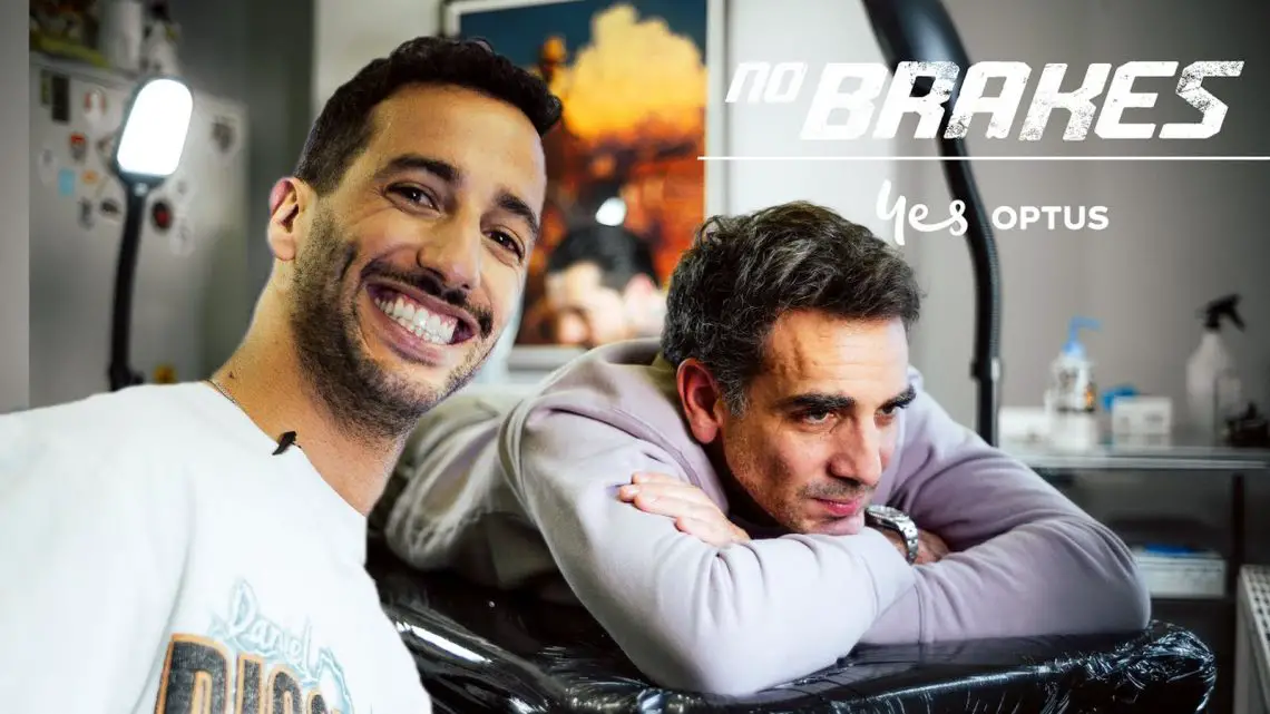 Twee mannen in een tattooshop: de één glimlachend naar de camera, de ander rustend op een armleuning. Achtergrond toont tattoo-apparatuur en wanddecoraties. De tekst luidt "geen remmen" en "ja OPTUS." Zou dat Daniel Ricciardo kunnen zijn die zijn avontuurlijke geest omarmt?