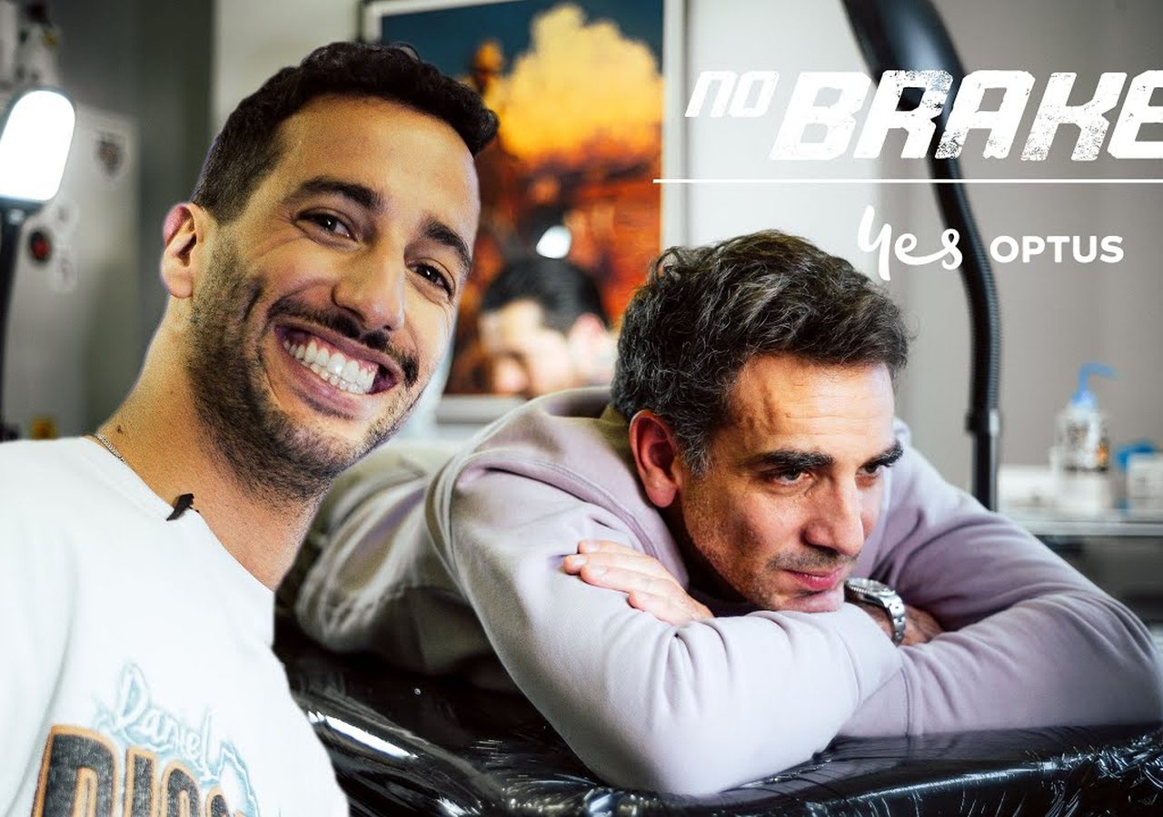 Twee mannen in een tattooshop: de één glimlachend naar de camera, de ander rustend op een armleuning. Achtergrond toont tattoo-apparatuur en wanddecoraties. De tekst luidt "geen remmen" en "ja OPTUS." Zou dat Daniel Ricciardo kunnen zijn die zijn avontuurlijke geest omarmt?