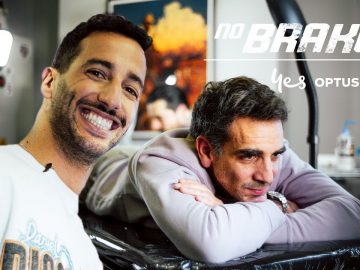 Twee mannen in een tattooshop: de één glimlachend naar de camera, de ander rustend op een armleuning. Achtergrond toont tattoo-apparatuur en wanddecoraties. De tekst luidt "geen remmen" en "ja OPTUS." Zou dat Daniel Ricciardo kunnen zijn die zijn avontuurlijke geest omarmt?