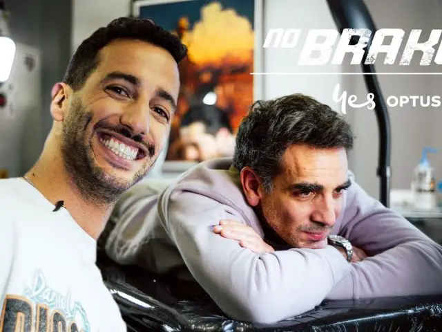 Twee mannen in een tattooshop: de één glimlachend naar de camera, de ander rustend op een armleuning. Achtergrond toont tattoo-apparatuur en wanddecoraties. De tekst luidt