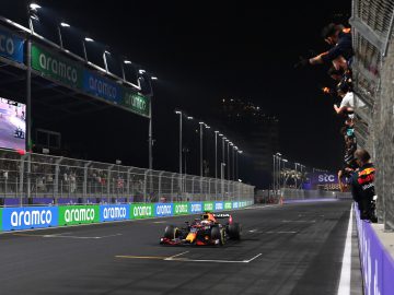 Een Formule 1-auto racet onder de nachtelijke hemel, terwijl toeschouwers van achter een hekwerk juichen. Tussen de snel rijdende voertuigen valt de auto van Max Verstappen op. Sponsorbanners zijn zichtbaar op de slagbomen, wat bijdraagt aan de opwindende sfeer.