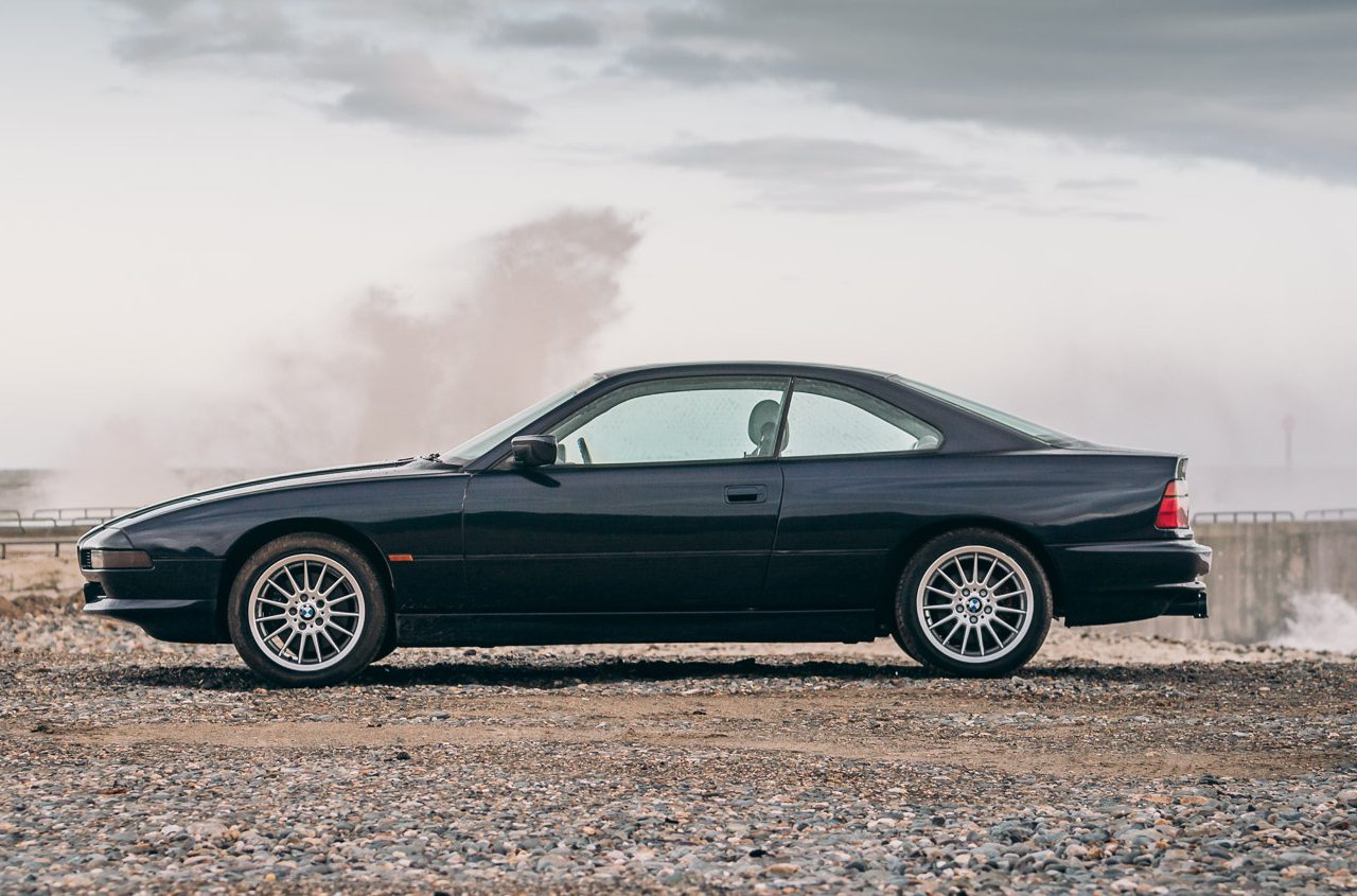 Een slanke BMW 8-serie coupé geparkeerd op een rotsachtige kust met oceaangolven op de achtergrond onder een bewolkte hemel.
