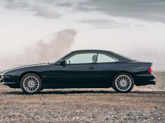 Een slanke BMW 8-serie coupé geparkeerd op een rotsachtige kust met oceaangolven op de achtergrond onder een bewolkte hemel.