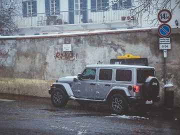 Een zilveren Jeep staat geparkeerd in een besneeuwde straat naast een muur versierd met graffiti. Borden met parkeerbeperkingen en snelheidsbeperkingen zijn duidelijk zichtbaar.