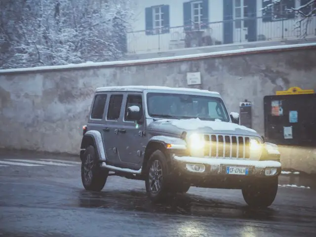 Een zilveren Jeep SUV rijdt op een natte en besneeuwde weg naast een stenen muur en gebouw. Er heeft zich sneeuw opgehoopt op het dak van het voertuig en de koplampen zijn ingeschakeld.