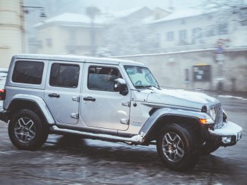 Een zilveren Jeep SUV die op een koude, mistige dag over een besneeuwde weg met gebouwen op de achtergrond navigeert.