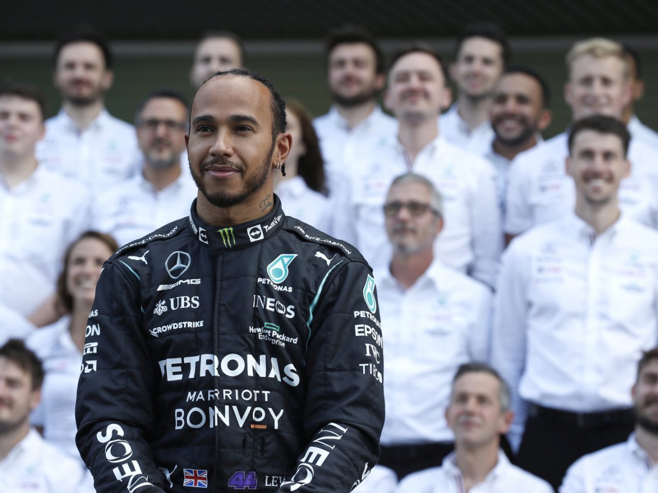 Een autocoureur in een zwart racepak staat voor een grote groep teamleden in witte shirts, die doen denken aan de iconische aanwezigheid van Lewis Hamilton op het circuit.