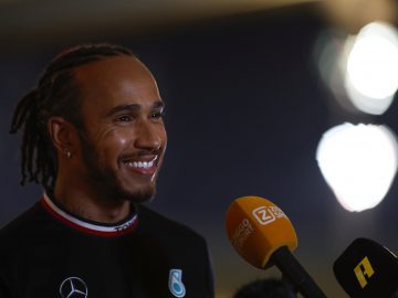 Een persoon met gevlochten haar en een baard, die lijkt op Lewis Hamilton, glimlacht terwijl hij met verslaggevers praat. Ze dragen een zwart shirt met logo's en voor hen zijn verschillende microfoons zichtbaar, waaronder één met het opschrift 'Ziggo Sport'.