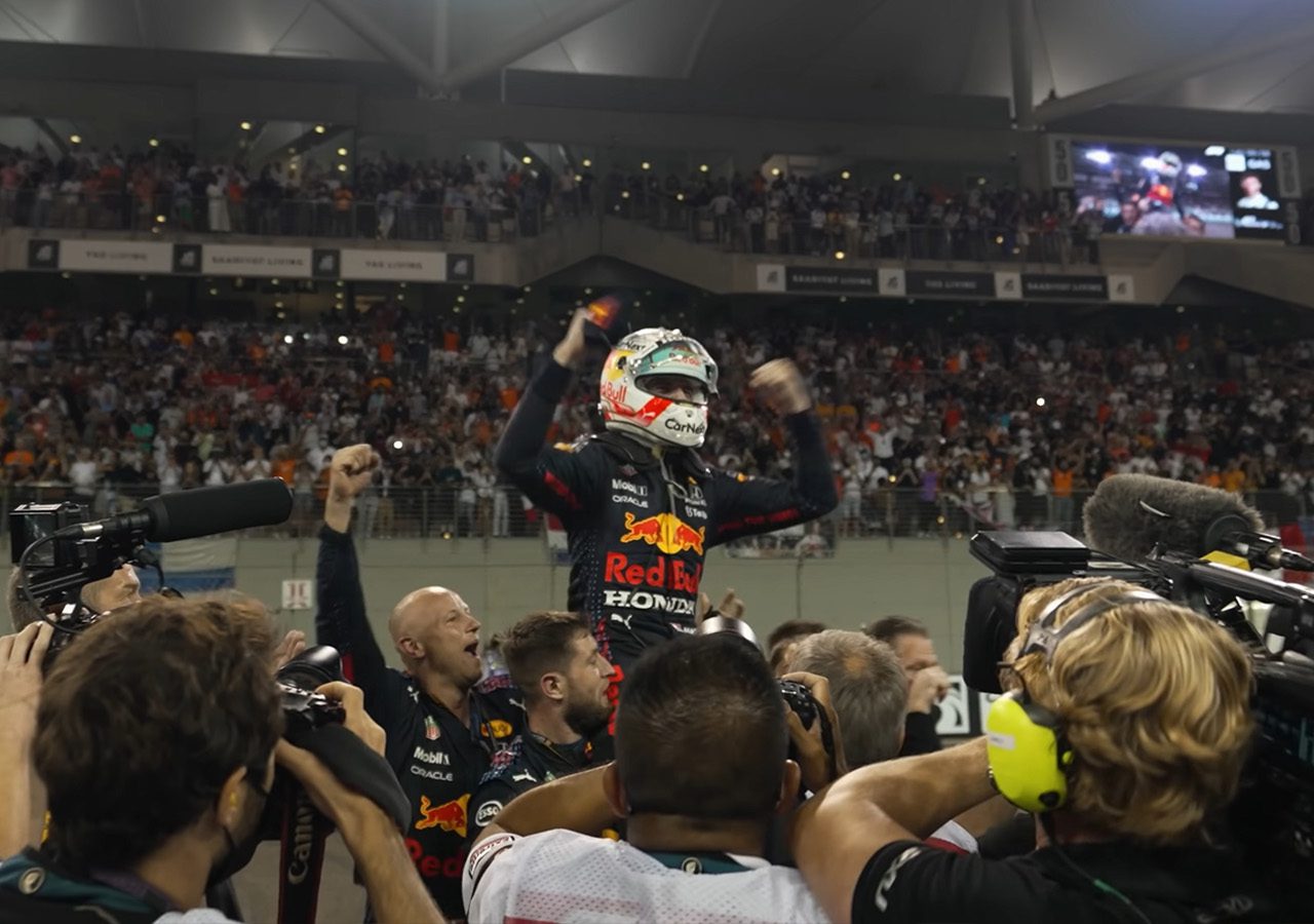 Max Verstappen, in Red Bull-kledij, viert feest met opgeheven armen te midden van een menigte fotografen en teamleden op een racecircuit.