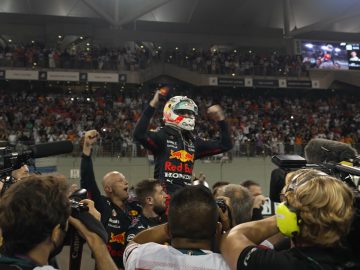 Max Verstappen, in Red Bull-kledij, viert feest met opgeheven armen te midden van een menigte fotografen en teamleden op een racecircuit.