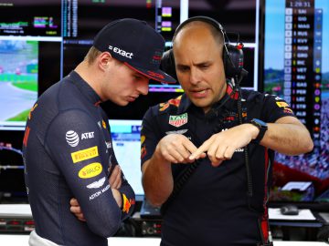 Twee mannen discussiëren terwijl ze voor monitoren staan die racegegevens weergeven. De één, die Max Verstappen lijkt te zijn, draagt een racepak en pet, terwijl de ander een koptelefoon draagt en naar iets voor zich wijst.