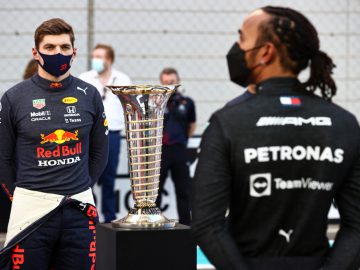 Twee van de best betaalde sporters van 2021 staan aan weerszijden van een grote trofee, de een naar voren gericht in een Red Bull Honda-racepak en de ander wegkijkend in een Petronas AMG-pak, met een hekwerk op de achtergrond.