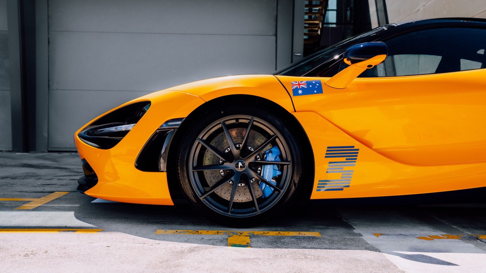 Close-up zijaanzicht van een gele sportwagen geparkeerd voor een gebouw, met een sticker van de Australische vlag op de zijkant. De afbeelding benadrukt het voorwiel en de strakke designkenmerken van de auto, die doen denken aan de gedurfde racestijl van Daniel Ricciardo.