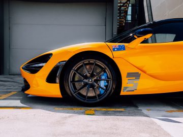 Close-up zijaanzicht van een gele sportwagen geparkeerd voor een gebouw, met een sticker van de Australische vlag op de zijkant. De afbeelding benadrukt het voorwiel en de strakke designkenmerken van de auto, die doen denken aan de gedurfde racestijl van Daniel Ricciardo.