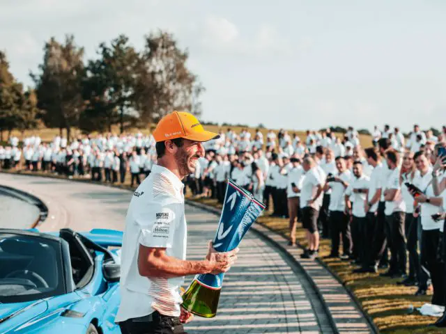 Een persoon met een oranje pet houdt een trofee vast naast een blauwe sportwagen, met op de achtergrond een menigte mensen in witte shirts. Het lijkt een overwinningsmoment voor Daniel Ricciardo.