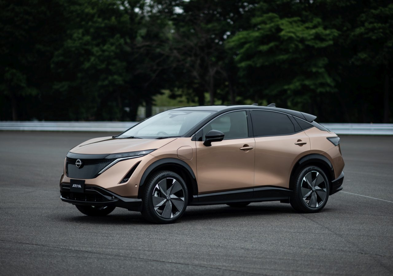 Een bronzen Nissan Ariya SUV staat geparkeerd op een verharde weg met groene bomen op de achtergrond.