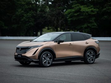 Een bronzen Nissan Ariya SUV staat geparkeerd op een verharde weg met groene bomen op de achtergrond.