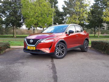 Een rode Nissan Qashqai crossover-SUV staat geparkeerd op een asfaltoppervlak in een parkachtig gebied met groene bomen op de achtergrond. Het voertuig beschikt over een geel kenteken met nummer L-185-VD.