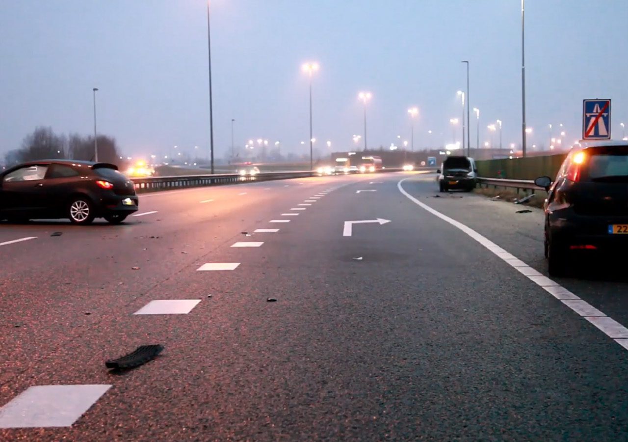 Een snelweg met weinig verkeer, waarop twee auto's te zien zijn die bij een ongeval betrokken zijn. Er is puin zichtbaar op de weg en beide auto's staan aan weerszijden van de rijbaan. De lucht is bewolkt, wat duidt op IJzelomstandigheden die mogelijk de aanvaring hebben veroorzaakt.