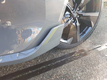 Close-up van de rechter voorband en lichtmetalen velg van een Peugeot 508 PSE op een kiezeloppervlak, waarbij de carrosserie nabijgelegen huizen weerspiegelt. Naast de auto loopt een klein afvoerkanaaltje over de grond.