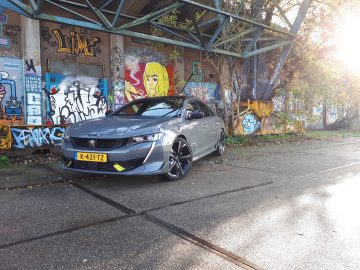 Een Peugeot 508 PSE met een gele kentekenplaat staat geparkeerd voor een met graffiti bedekte muur onder een metalen structuur, waarbij overwoekerde vegetatie bijdraagt aan de sfeer terwijl zonlicht schaduwen werpt.