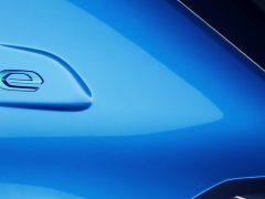 Close-up van de carrosserie van een blauwe elektrische auto met een 'e'-logo in reliëf aan de achterkant. Het ontwerp is strak met reflecterende oppervlakken.