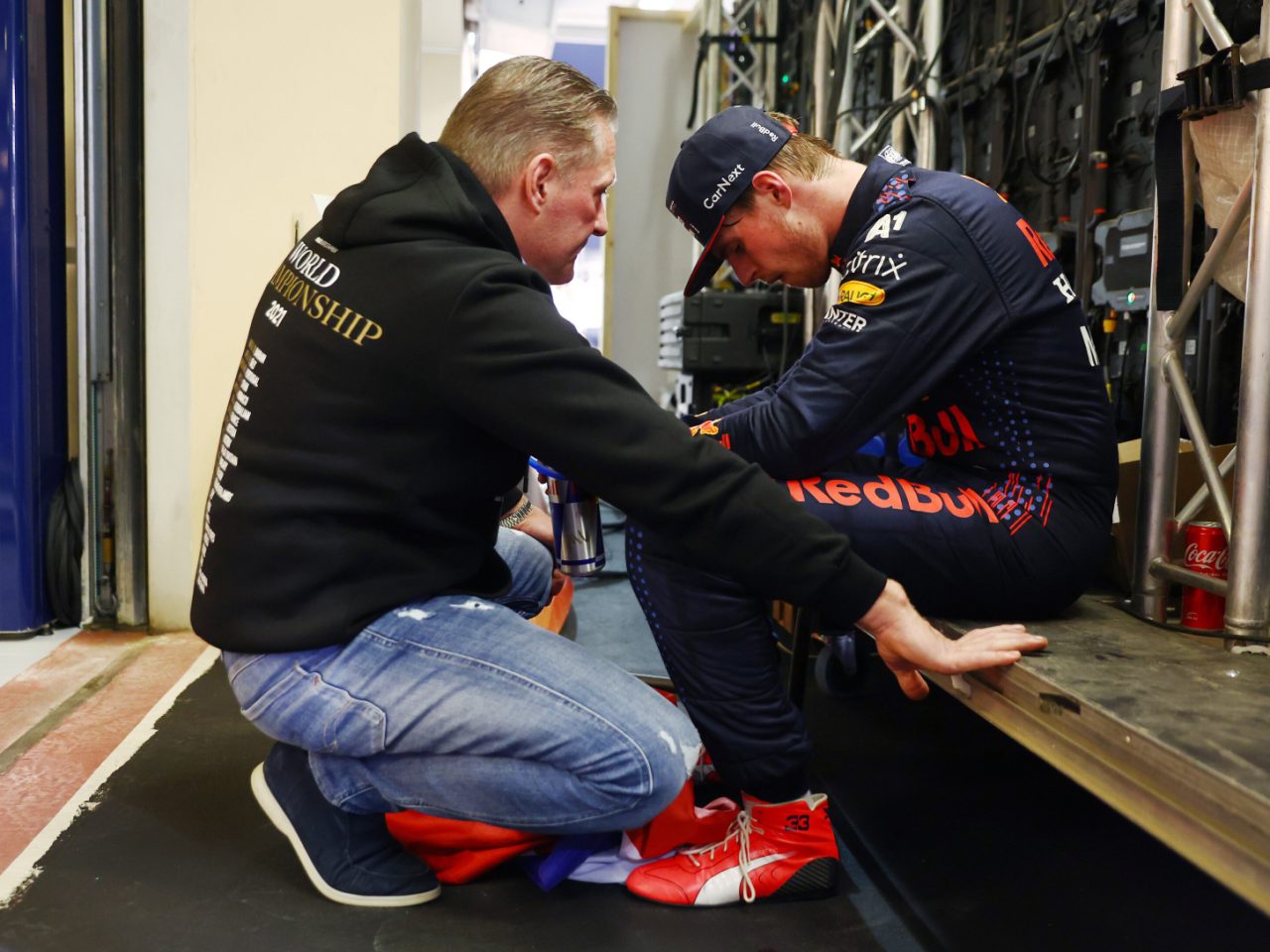 Een persoon in een zwarte hoodie knielt en spreekt met een zittende Max Verstappen, die in zijn racekleding aandachtig lijkt te luisteren. Beide individuen bevinden zich in een backstage-ruimte met technische apparatuur in de buurt.