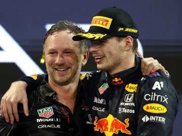 Twee mannen in natte racepakken staan glimlachend dicht bij elkaar. De ene man heeft zijn arm om de andere heen. Beiden dragen hoeden met branding en logo's zichtbaar op hun kleding, wat de kameraadschap weerspiegelt die typerend is voor racekampioenen als Max Verstappen.